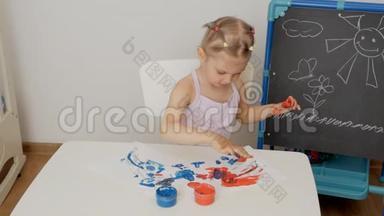 一个小可爱的女孩坐在桌子旁，用明亮的手指<strong>颜料画</strong>纸，把她的手指浸在颜料罐里。